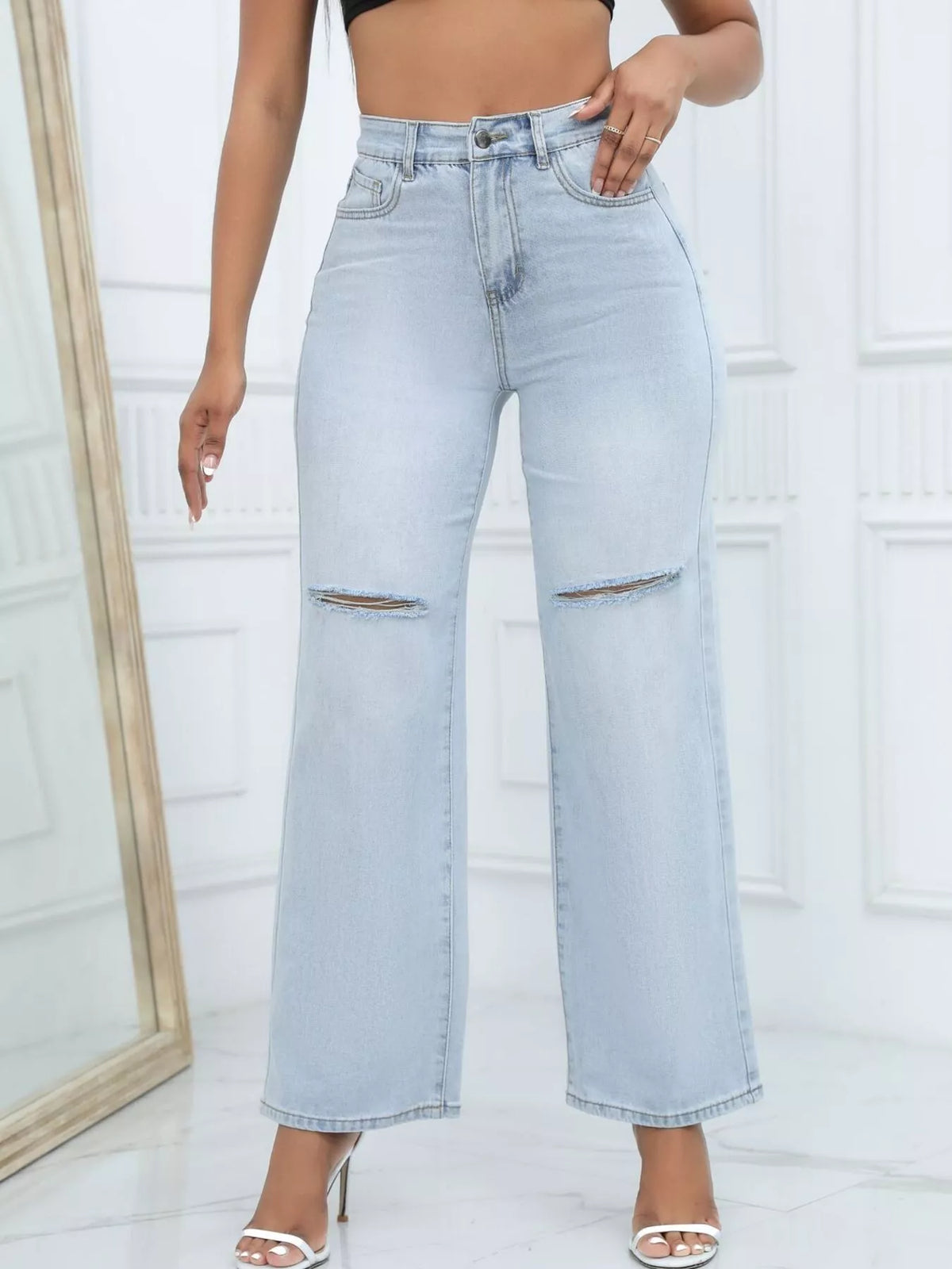 A model wearing light-wash blue high-waisted wide-leg jeans with distressed rips on the thighs, paired with white heeled sandals in a bright white room.