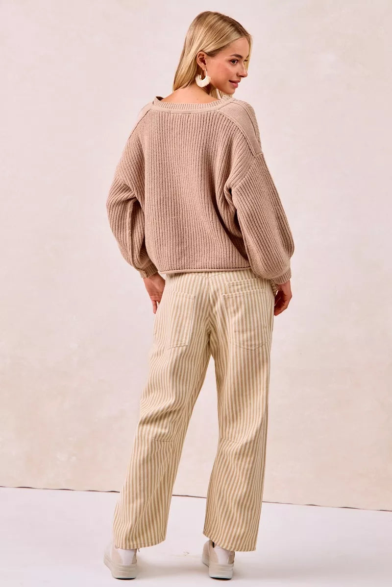 Woman wearing a beige ribbed sweater and striped pants against a plain background