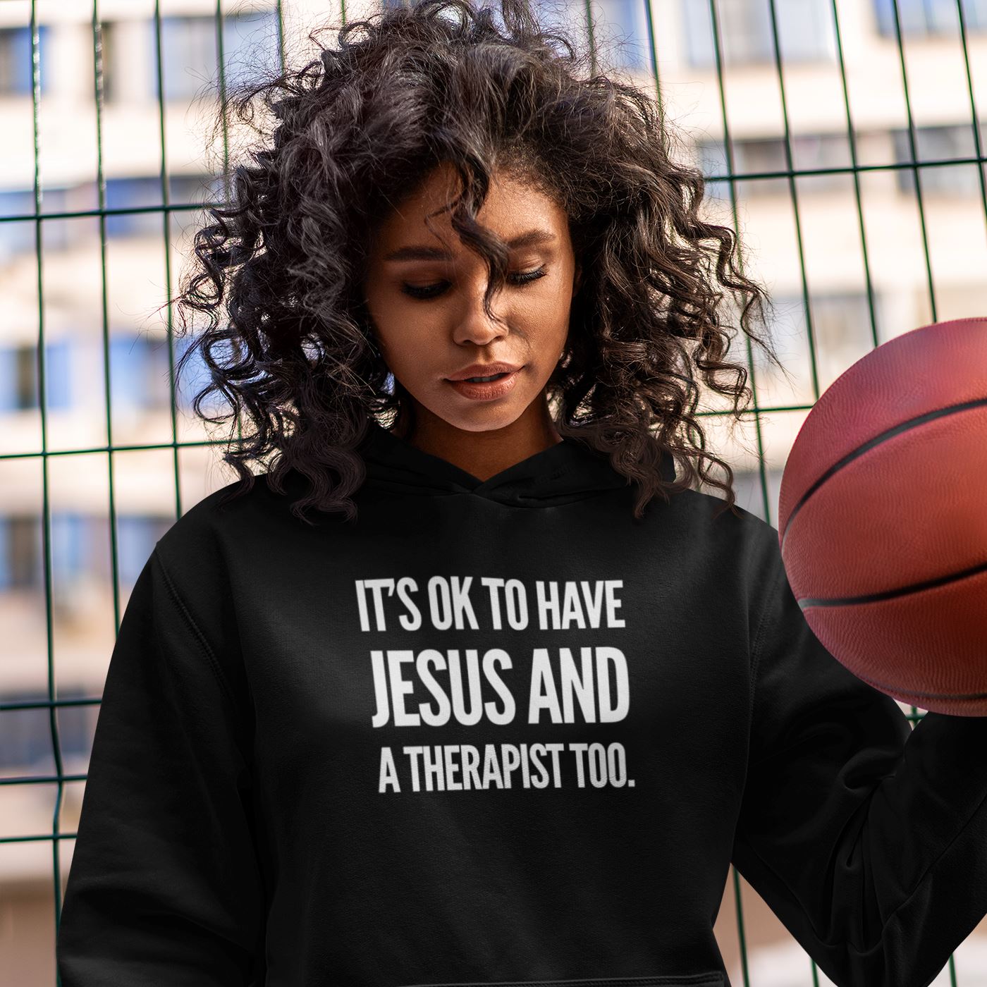 Person wearing a black hoodie with text holding a basketball, standing against a wire fence.