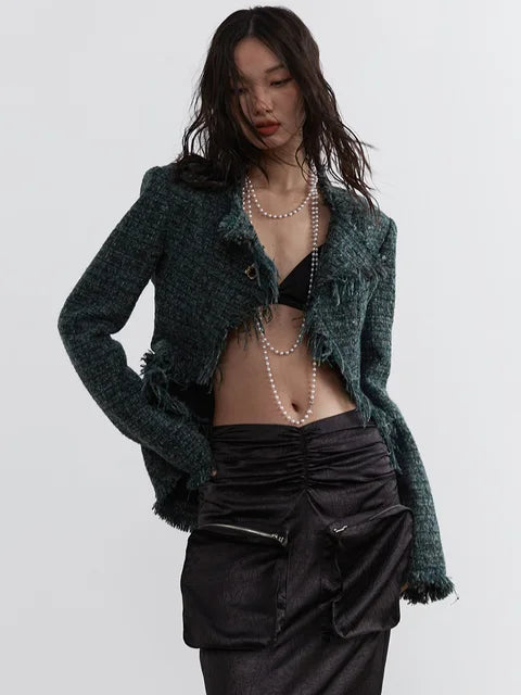 A person wearing a green frayed tweed jacket with a V-neck collar and pockets, paired with a black outfit. The jacket has a open front and the person is also wearing a necklace.