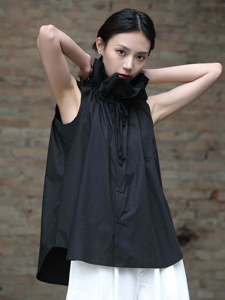 A woman modeling a black ruffle collar blouse with sleeveless design, standing against a brick wall background.