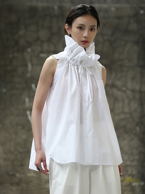 A woman modeling a white sleeveless blouse with a ruffle collar, made of polyester, against a concrete background.