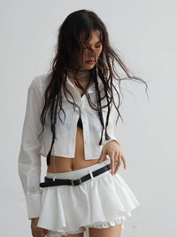A person wearing a white button-up blouse with a mini skirt, accessorized with a belt, necklace, and earrings.