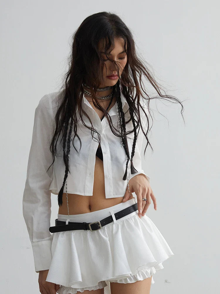 A person wearing a white button-up blouse with a mini skirt, accessorized with a belt, necklace, and earrings.