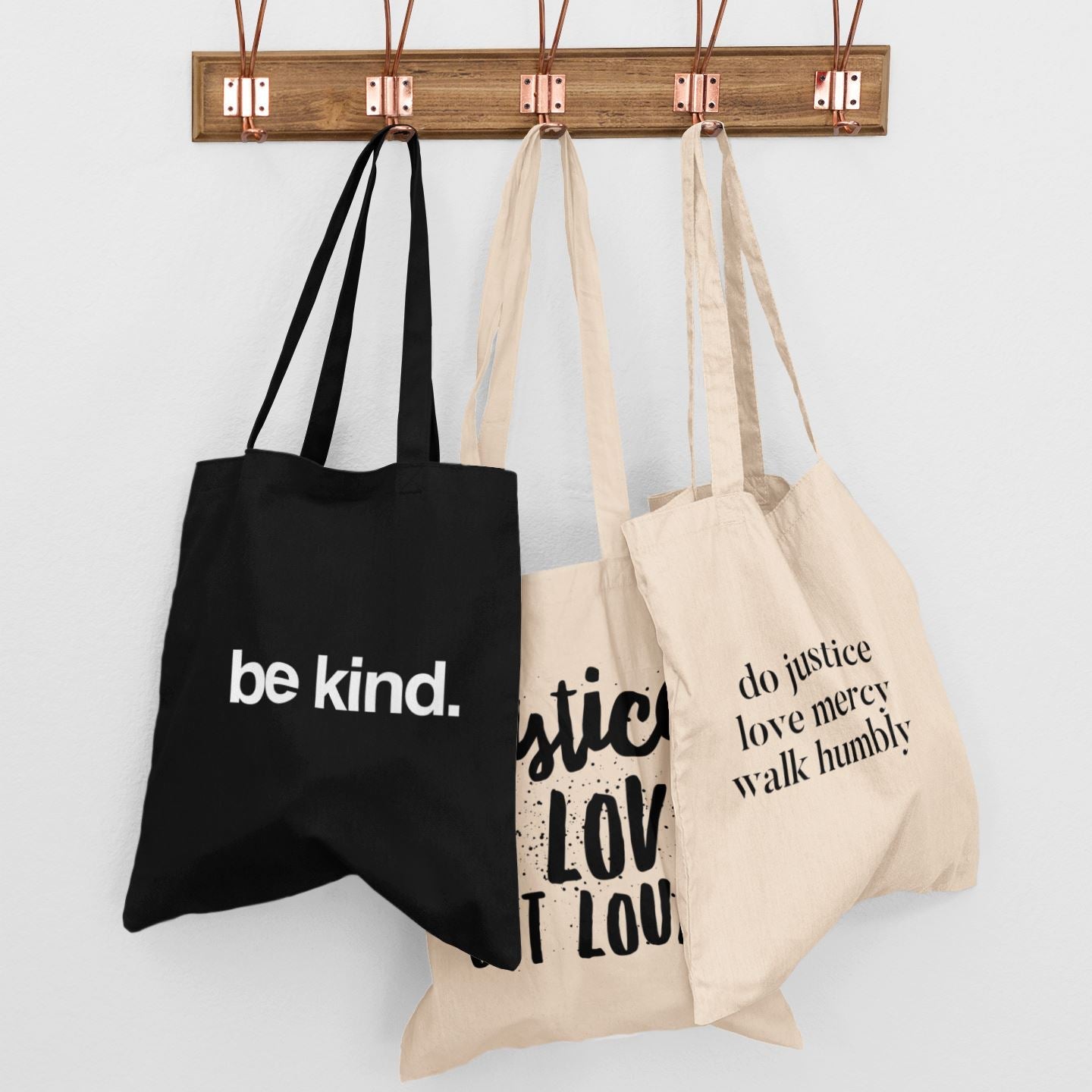 Three tote bags with motivational phrases hanging on a wooden rack against a white background.