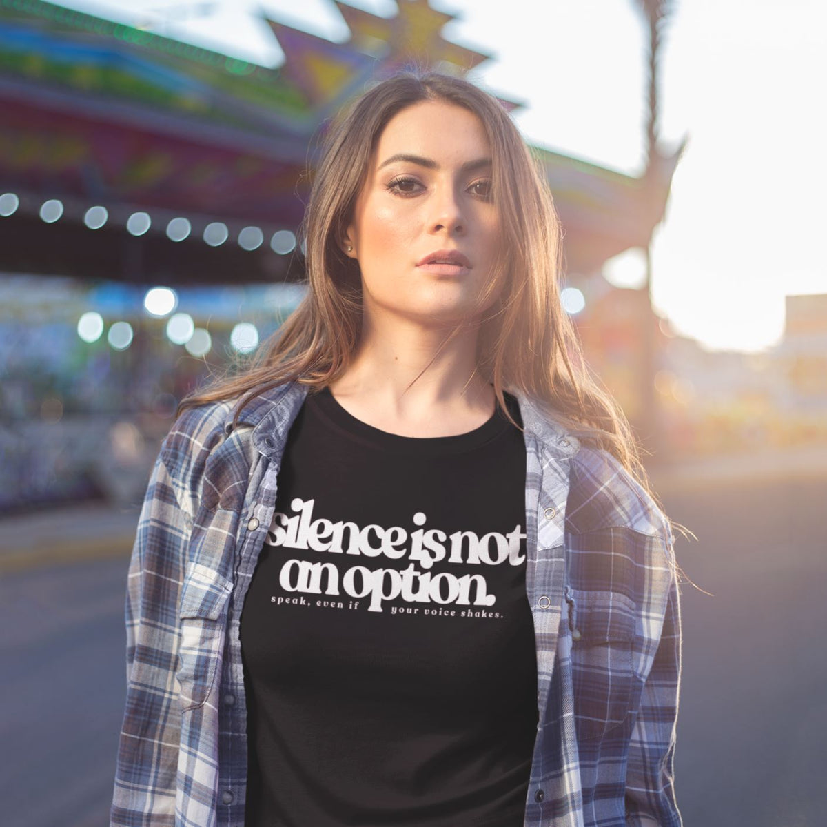 Woman wearing a black t-shirt with 'silence is not an option' text in front of a blurred amusement park background.