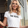Woman wearing a white t-shirt with 'be kind.' text, standing outdoors.