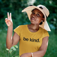 Woman wearing a yellow 'be kind.' shirt with a wide-brimmed hat outdoors.