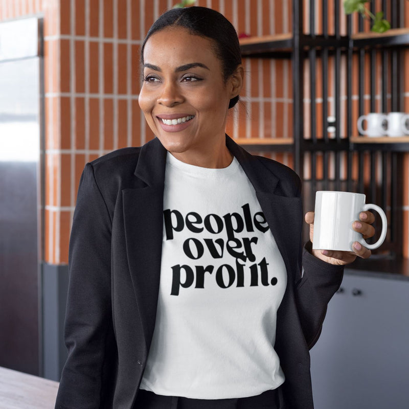 Woman wearing a shirt with 'people over profit' text, holding a mug in a kitchen setting.