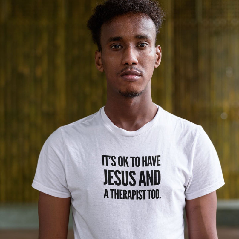 Person wearing a white t-shirt with text 'It's ok to have Jesus and a therapist too.' against a blurred background.
