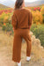 Model standing on the side of a country dirt road overlooking ridge with trees showing the back of Journey Pant- color is brown